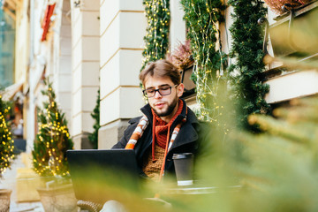 Elegant beard entrepreneur in glasses working with a laptop outdoors sitting in the street cafe in winter time.