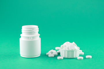 Close up white pill bottle with spilled out pills and capsules in cap on emerald green background with copy space. Focus on foreground, soft bokeh. Pharmacy drugstore concept