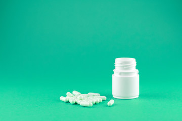 Close up white pill bottle with spilled out capsules on emerald green background with copy space. Focus on foreground, soft bokeh. Pharmacy drugstore concept