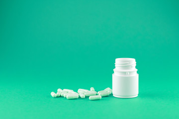 Close up white pill bottle with spilled out capsules on emerald green background with copy space....