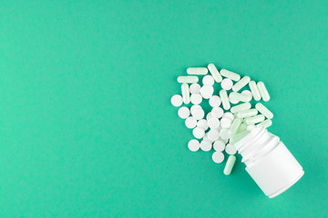 Close up white pill bottle with spilled out pills and capsules on emerald green background with copy space. Focus on foreground, soft bokeh. Pharmacy drugstore concept. Top view