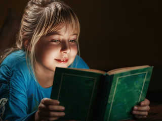 a little blonde girl in a blue blouse lies in a dark room and reads a green book