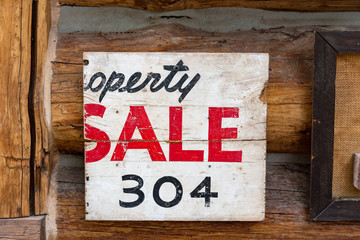 An old broken property for sale sign with vintage phone number hanging on a log house wall. The number on the sign is 304.