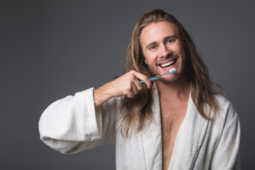 Healthy smile. Waist up portrait of laughing longhaired guy brushing his teeth. Isolated on grey background