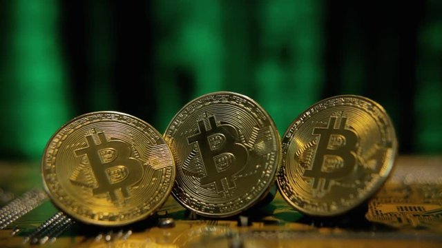 A trio of physical golden bitcoins (digital virtual crypto-currency) on a printed circuit board. Background: blurred glowing green matrix source code rain falling down.
