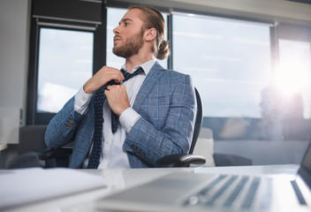 Low angle of tranquil office worker sitting at the table. He is straightening a tie and looking aside