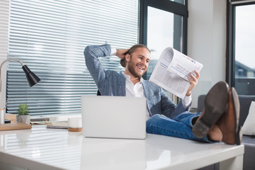 Smiling guy relaxing in the office with mug of beverage. He is reading newspaper with happiness