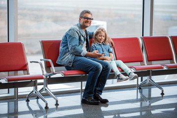 Enjoying rhythm. Full length of delighted middle-aged father and little daughter are sitting on airport bench while waiting for boarding. They are listening to music on smartphone through earphones