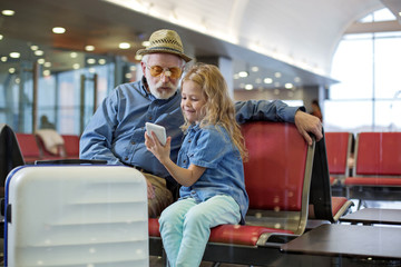 Look at this. Optimistic granddaughter is sitting on red bench at airport lounge with her...