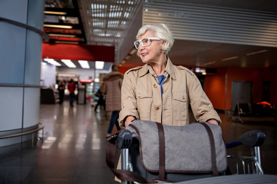 Positive senior charming woman in glasses is standing in modern terminal with airport trolley loaded with luggage. She is looking aside with smile. Copy space in the left side