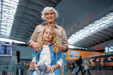 Obraz na płótnie Canvas Feeling wonderful. Low angle portrait of cheerful old granny is standing behind her little grandchild and hugging her. They are expressing gladness while waiting for flight at terminal lounge