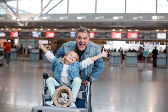 Happy life. Portrait of joyful mature man is pushing airport trolley with suitcases and his nice little daughter who is sitting on it while rising her hands up. Male is looking at camera with joy