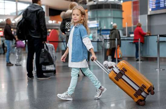 Favorite holiday. Full length portrait of optimistic trendy little girl is walking along airport lounge while carrying orange suitcase. She is looking at camera with slight smile. People in background