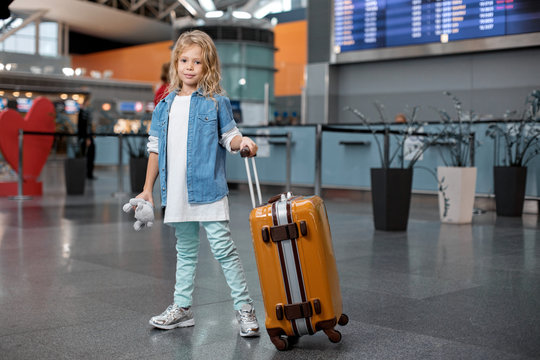 First travel. Full length portrait of positive stylish cute blond child is standing at airport building and carrying her suitcase while keeping toy bear. She is looking at camera with joy. Copy space