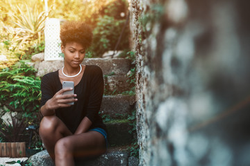 Admirable young Brazilian female is sitting on the stone step of staircase and taking selfie using camera of her mobile telephone, with copy space place for advertising, text message or your logo - Powered by Adobe