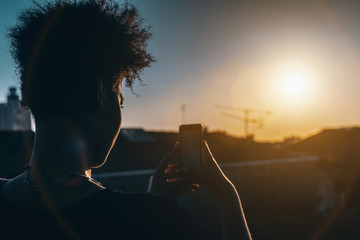 Rear view of black curly woman taking pictures of stunning sunset on her smartphone; young Brazilian female is photo shooting urban sundown on cellphone to post into her social network accounts - Powered by Adobe