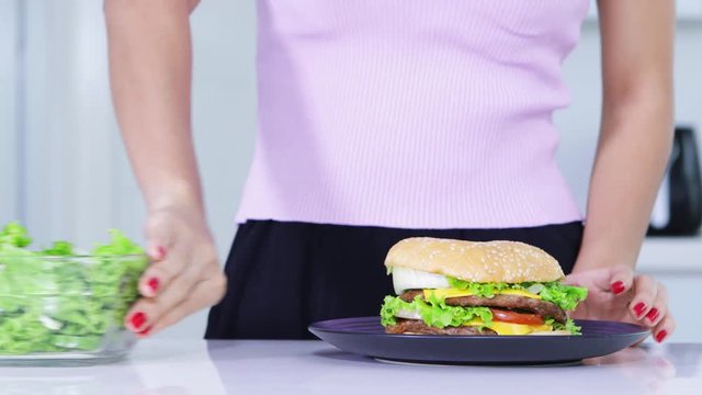 Dieting concept. Close up of unknown woman hands refuse a bowl of salad and choosing to eat a plate of burger. Shot in 4k resolution
