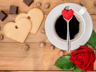 The arrangement for Valentines day of a cup of coffee with a heart shape paper on metal tea spoon and red rose and heart shape candy on wooden slab