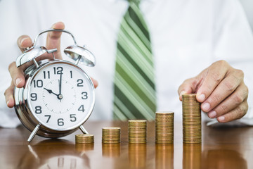 Businessman hand holding money coin stack arranged as a graph with alarm clock on wooden table, concept of time to money growth and saving money
