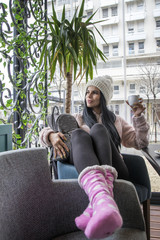 Girl with long dark hair and cap sitting on cafe and drink coffee, she wearing fluffy jacket and small backpack beside her