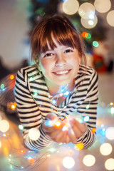 Fototapeta na wymiar Young girl holds light colorful garland in hands before Christmas Tree