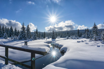 Winter mountain landscape with stream and wooden bridge. Trees and the sun are reflected in the...