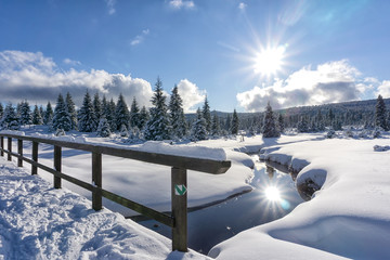 Winter mountain landscape with stream and wooden bridge. Trees and the sun are reflected in the water. Izera Mountains, Czech Republic, Europe.