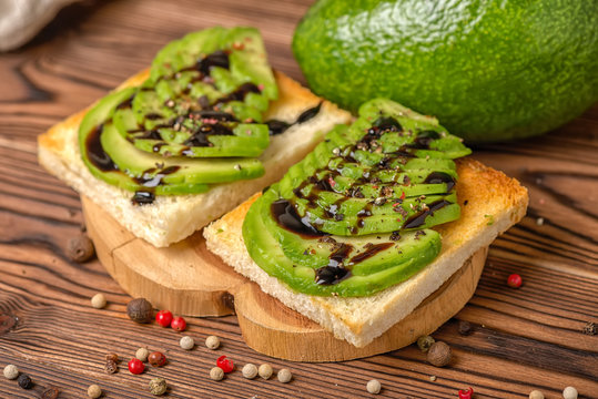 made sandwiches with sliced avocado, white bread toast decorated alligator pear fruit, drizzled sauce and sprinkle pepper on wooden background, vegetarian food