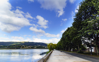 Fototapeta premium Wide angle photo of the promenade in the bay of Pontedeume in La Coruna, Spain with mountains with houses and a blue sky with white clouds