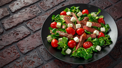 Juicy Beef Sirloin Steak Salad with roasted tomatoes, feta cheese and green vegetables in a black plate. healthy food