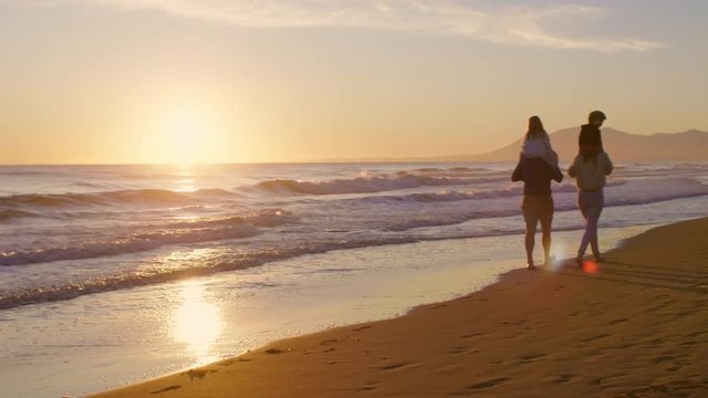 Young family walking and playing on beach with children on shoulders in sunset
