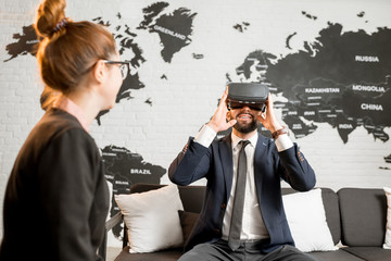 Businessman using virtual reality glasses sitting at the travel agency office with woman manager and world map on the background