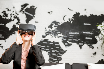 Young businesswoman trying virtual reality glasses sitting indoors with world map on the background