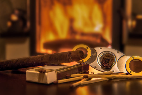 Time to enjoy! Quality cigars lie on a wooden table and other accessories such as cigar cutters and matches. In the background a fireplace. Focus on the cigars. Concept: life style or health