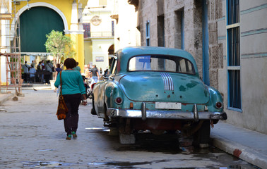 Straßenszene, türkis grüner Oldtimer ohne Räder auf Steinen aufgebockt, Frau im türkis grünem Oberteil und Umhängetasche läuft über die Kopfsteinpflaster Straße in Havanna auf Kuba