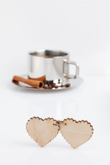 Two wooden hearts connected by a twine on a white table. At the back is a cup of coffee with grains and a cinnamon stick.