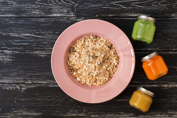 Muesli in a pink plate, kiwi juice, carrot juice, apple puree, wooden dark background. View from above. space for text.
