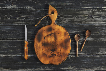Round old cutting board with a wooden spoon and a knife on a dark rustic background. View from above.