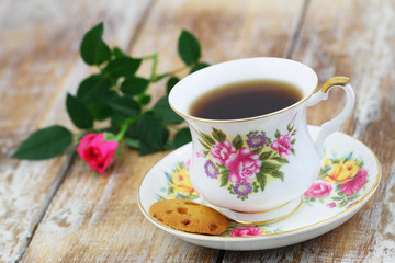 Tea in vintage porcelain cup, crunchy cookie and pink wild rose on rustic wooden surface
