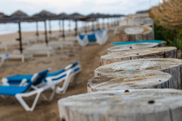 cloesup of wooden poles, standing at the beach, dividing the shore from the land. Sun umbrellas and beach loungers stand on the blurry beach in the background.