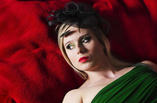 portrait of a blonde woman wearing makeup and a black retro hat lying on a red blanket