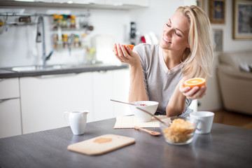 Obraz na płótnie Canvas Beautiful blond caucasian woman posing in her kitchen, while drinking coffee or tea and eating a healthy breakfast meal full of cereal and other healthy foods, including fruit