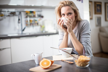 Beautiful blond  caucasian woman posing in her kitchen, while drinking coffee or tea and eating a healthy breakfast meal full of cereal and other healthy foods, including fruit