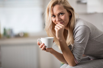 Beautiful blond  caucasian woman posing in her kitchen, while drinking coffee or tea and eating a healthy breakfast meal full of cereal and other healthy foods, including fruit