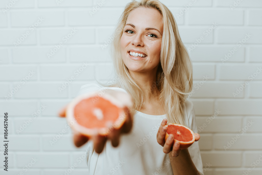 Wall mural Beautiful blond  caucasian woman posing in front of a white brick backround and  having fun with a set of two red oranges