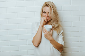 Beautiful blond  caucasian woman posing in front of a white brick backround and  having fun with a cup or two cups of coffee, maybe tea