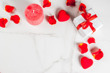 Valentine's day background with rose flower petals, white wrapped gift box with red ribbon and holiday red candle, on white marble background, copy space top view