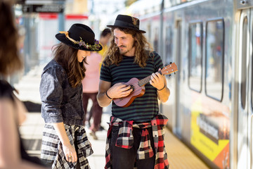 Two happy young free spirit travelers play guitar and sing waiting for public transportation 