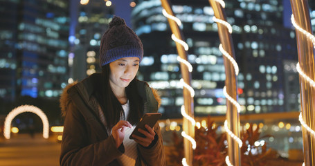 Woman use of smart phone in the city at night, surf online on smart phone with urban city background, woman wearing winter jacket