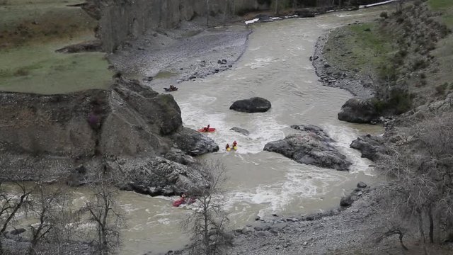 Four pairs of people run down the wild river on the twin-hull boats and drifting the turn. The kayaks goes to the stones in the middle of the race and people uses the paddles to ride aside. The gray
